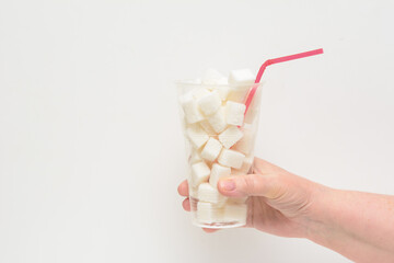 In a woman's hand, a plastic glass filled with lumps of sugar. In a glass there are red straws. White background. Concept - sugar-free diet. Copy space