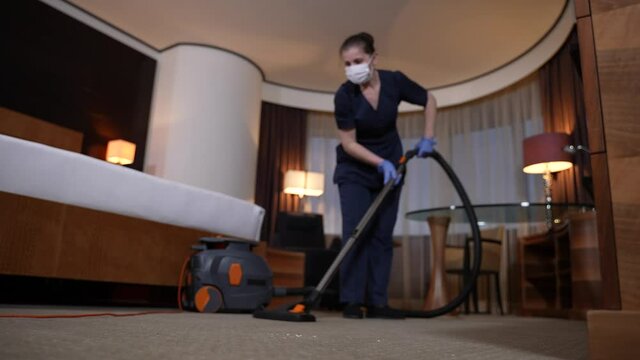 Responsible hotel staff using vacuum cleaner while serving room. Maid in uniform, protective mask and gloves working with vaccum cleaner while cleaning hotel room during covid-19 epidemic spread