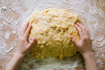 a child kneads dough for a cake with his hands on a wooden table sprinkled with flour, top view