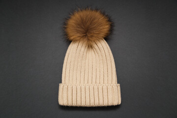 Knitted beige hat with fur pompon on gray background. Cozy knitted accessory in trendy colors, warm concept