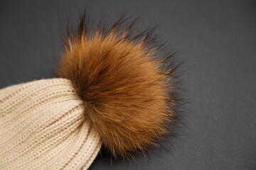 Knitted beige hat with fur pompon on gray background. Cozy knitted accessory in trendy colors, warm concept