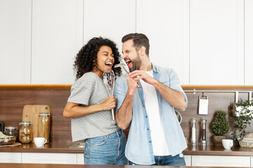 Happy interracial couple dancing in the kitchen, singing while cooking breakfast or dinner, young African American woman and a handsome man smiling and having fun together, holding kitchen utensils
