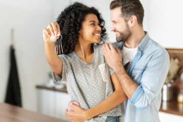 Happy young interracial married couple holding a key ring in hand, hugging and looking at each other with love, standing in the modern kitchen of their new home, excited owners of a family apartment