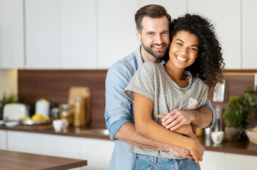 Smiling man hugging from behind charming African American woman, two people standing and joyfully looking at camera. Young international couple happily spending time in cozy modern kitchen at home. - Powered by Adobe