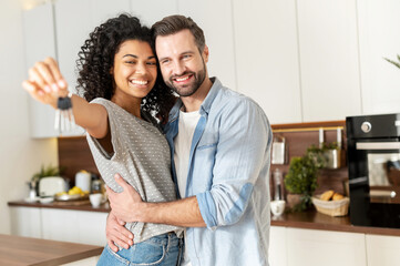 Fototapeta na wymiar Young interracial married couple homeowners smiling, showing keys from a new apartment, hugging and looking at the camera, standing in the kitchen and celebrating moving in a new home, family concept