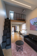Stylish, modern interior of a teen's living room in a private house.Work space with white desk,chair. Black leather sofa, round table for relaxation. Sleeping area with a bed at the top of the stairs