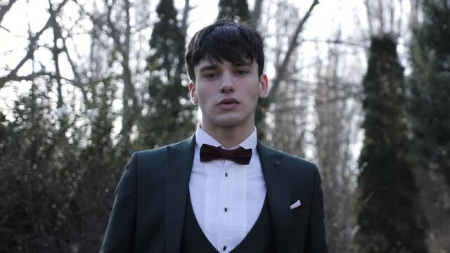 Confident young guy wearing stylish black suit set with white shirt and bow tie stands in autumn city park closeup slow motion