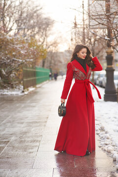 Long-haired woman in red maxi coat and stands near exquisite building