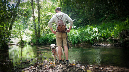 Hombre joven con mochila de piel y pantalón corto junto a río en bosque de Asturias con su perro...
