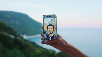 Close-up of hand holding smartphone during video call with man in front of the sea. Beautiful landscape in the background.