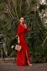 Stunning woman in red evening maxi dress posing in greenhouse