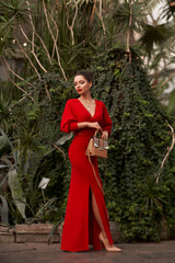 Stunning woman in red evening maxi dress posing in greenhouse