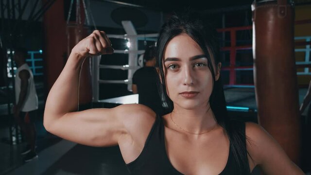 Attractive strong female athlete looking positive at camera boasting of muscles showing biceps in the gym. Boxing ring on background.