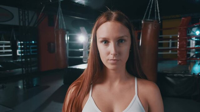 Attractive ginger caucasian girl professional fitness coach posing into camera at boxing ring. Boxing club. People training. Workout lifestyle.