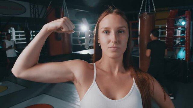 Portrait of nice strong young female showing bicep muscle smiling at camera. Bodybuilder lifestyle. Sportswoman. Boxing club.