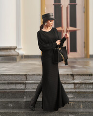 Elegant woman in stylish black maxi dress and cap posing outside exquisite building
