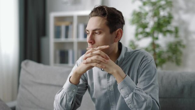 Pensive man in casual clothes sitting on grey couch and making important decision. Young guy lost in thoughts while staying at home.