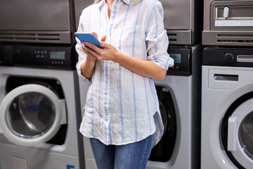 Woman stands next to the washing machine using mobile phone, waiting end of washing. cropped unrecognizable female in washing house