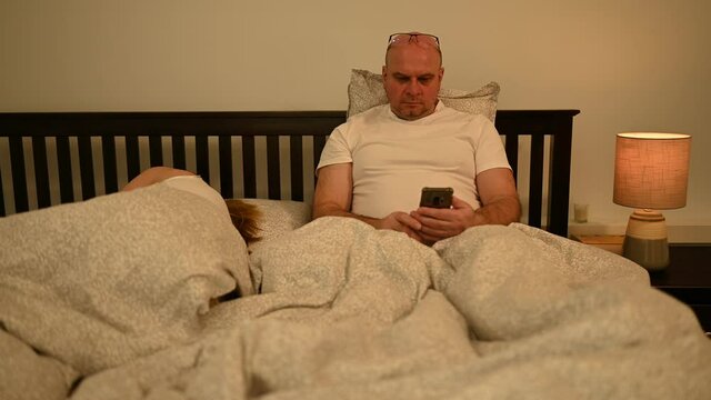 Real Husband And Wife In Their Early 50s In Bed At Home, While The Husband Is Getting Extremely Excited About The Shocking News, Message Or Information That He Shows To His Wife.