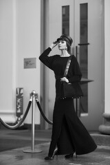 Elegant woman in stylish black maxi dress and cap posing outside exquisite building