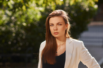 Young woman in black shirt, jeans and white blazer walking and posing outdoors on a summer day. Sun...