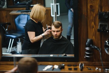 Professional hairdresser woman doing styling of handsome guy with electric hair clipper at barber shop