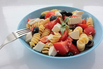 Itlian fusilli pasta salad with olives, tomato, and  feta cheese in blue bowl on white table background. Overhead view