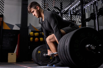 Fototapeta na wymiar Athlete standing on his knee preparing to make deadlift at gym, young caucasian man in black sportswear engaged in bodybuilding, concentrated on weightlifting. sport, cross fit concept
