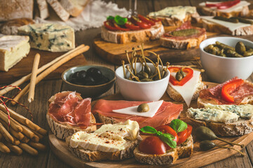 Baguette bread slices with cheese, bacon, salami, olives and capers as a starter, tapas, antipasti, mediterranean style on a rustic wooden table