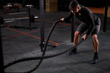 Fototapeta na wymiar Fit athlete doing battle rope exercise at cross fit gym, having intense training alone. Concentrated caucasian man doing cross fit exercise while working out in gym, in sportswear.