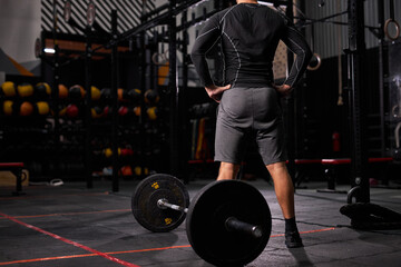 Fototapeta na wymiar rear view on sportsman standing near barbell taking break, rest. male in sportive outfit in modern gym