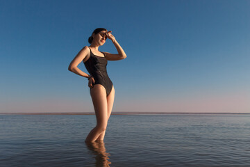 Summer vacation concept. Young beautiful slim woman wearing a black swimsuit stands in the sea and watching sunset. Copy space
