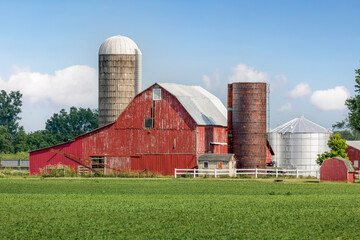 On the other side of a field of crops, an old red barn is surrounded by silos and other buildings and agricultural equipment on a rural Ohio farm in the American Midwest. - 411980989