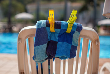 Beach towel fastened to plastic beach chair with pegs, sun shines blurred pool background