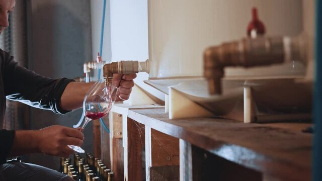 Side view of caucasian professional winemaker in apron pouring fermented fresh wine into glass. Winemaking facrory worker tasting wine in cellar.