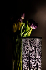 yellow and purple tulips in a vase behind a lamp with a forest motif