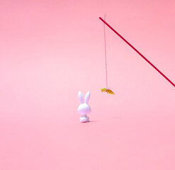 A toy rabbit sitting and staring in carrot on the stick with pastel pink background. Photo of its...