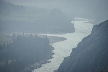 Misty mountain landscape with wide mountain river. Dark green gloomy scenery with confluence of two...