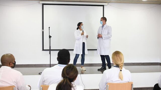 Two doctores wearing protective face mask to prevent viral infections standing with microphone on conference room stage, speaking to medical workers. High quality FullHD footage