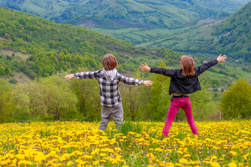 Boy, girl arms outstretched joyfully jumps on a meadow of dandelions in spring. Rest in nature, country recreation. Children having fun in the mountains together. Springtime. Joy and fun. Friendship