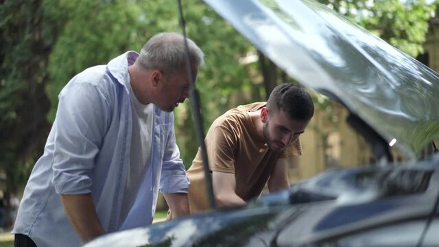 Focused young man examining engine in open car hood with middle aged guy. Caucasian son helping father fixing broken automobile. Transportation and support.