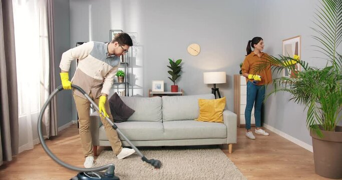 Young handsome happy man vacuuming carpet floor with vacuum cleaner in living room while his pretty Caucasian wife chatting and cleaning disinfecting picture, domestic work, housekeeping concept
