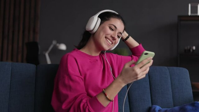 Attractive young woman listening to music in modern white headphone and use smartphone. slow motion. Beautiful girl enjoying music on couch at home. Female enjoys listening audio sound relaxing.