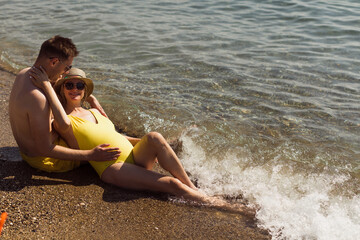 The lovely young pregnant couple is sitting by the ocean and chilling out.