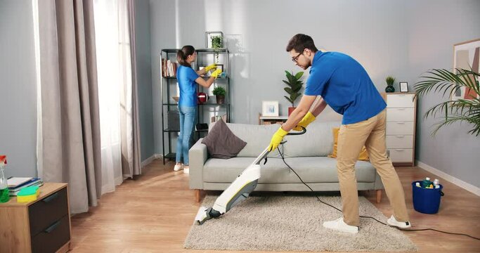 Young handsome happy professional man cleaner vacuuming carpet floor with vacuum cleaner in living room while his pretty Caucasian female colleague cleaning and disinfecting stuff, service concept