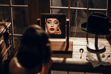 Obraz premium Cute girl looks in the mirror and does makeup, hair and furniture in retro style with vintage hats. A woman is going on a date or to work. Free space for text. Sixties and people in those days