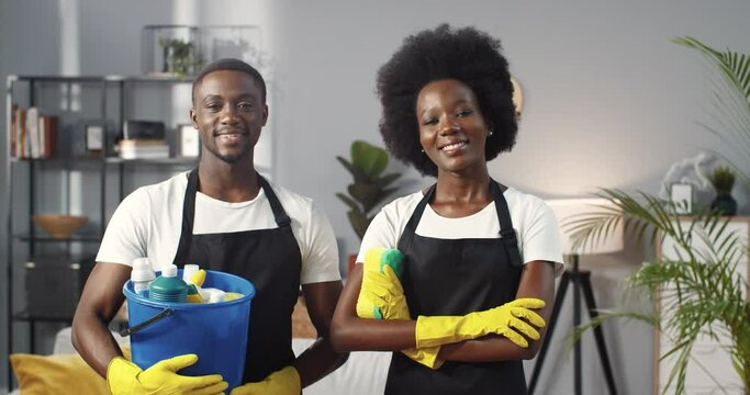 Portrait of African American joyful young couple team of professional cleaners in black aprons looking at camera and smiling holding bucket with cleaning products and sprays, cleaning business concept