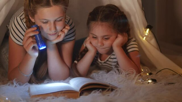 children in a wigwam tent play at night with a flashlight and read a book. happy family kid dream concept. children reading a book with a flashlight. kid dream reading a book in a stay home tent house