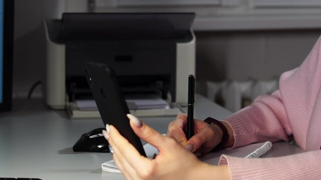 Close-up of muslim woman at office holding smartphone in her hand and speaking by videocall. Lady in hijab makes notes and smiling. She is writing essential points. Concept of professional occupation.