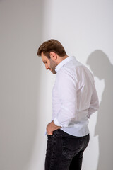A man in a white shirt, dark jeans and black boots stands sideways against a plain background....